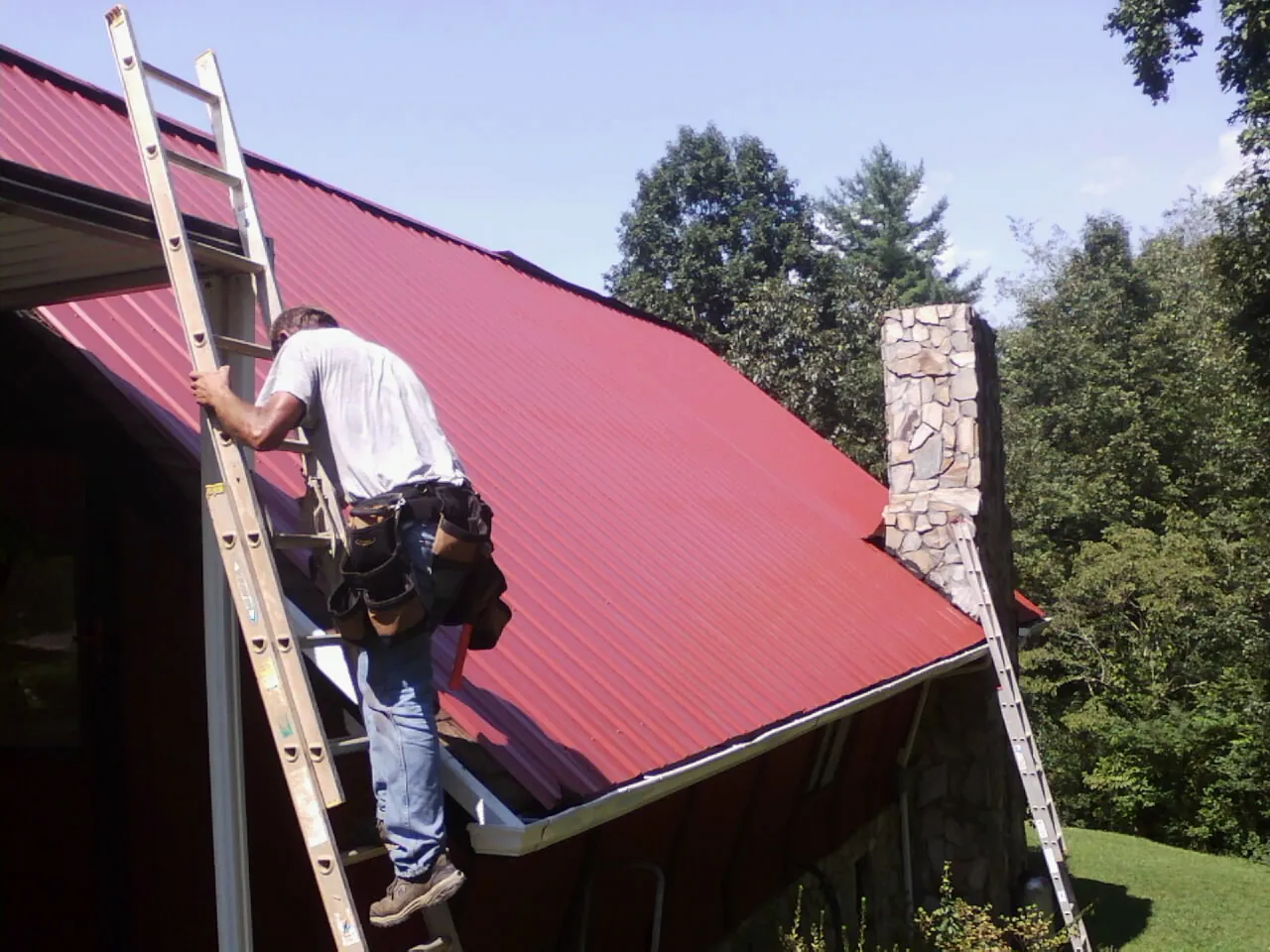 Roofer on a ladder beside a red metal roof and stone chimney — Soffit & Fascia Repair by Roofing Brothers in Pinebluff